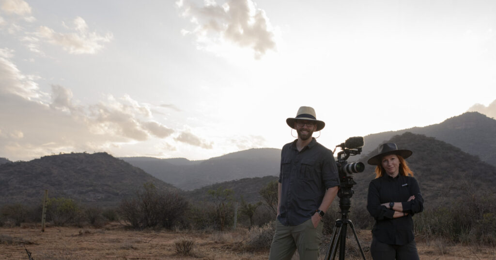 Two filmmakers with a camera on a tripod in a dry Kenyan landscape, mountains in the background at sunset