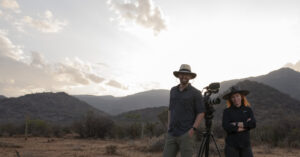 Two filmmakers with a camera on a tripod in a dry Kenyan landscape, mountains in the background at sunset